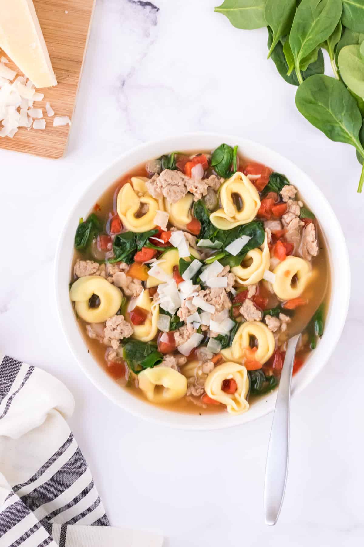 Bowl of Italian sausage tortellini soup with tomatoes, spinach and shredded Parmesan cheese with fresh spinach and block of cheese in upper background and black and white napkin in foreground.