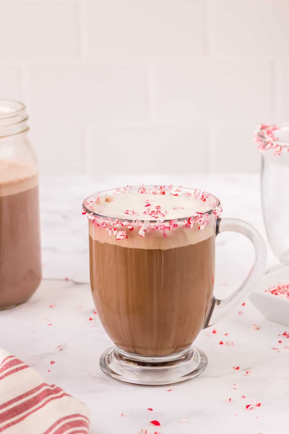 Glass mug with peppermint mocha coffee with crushed peppermint candies around lip of mug and extra mug in background and jar of creamer in left background with red and white striped linen in foreground. 