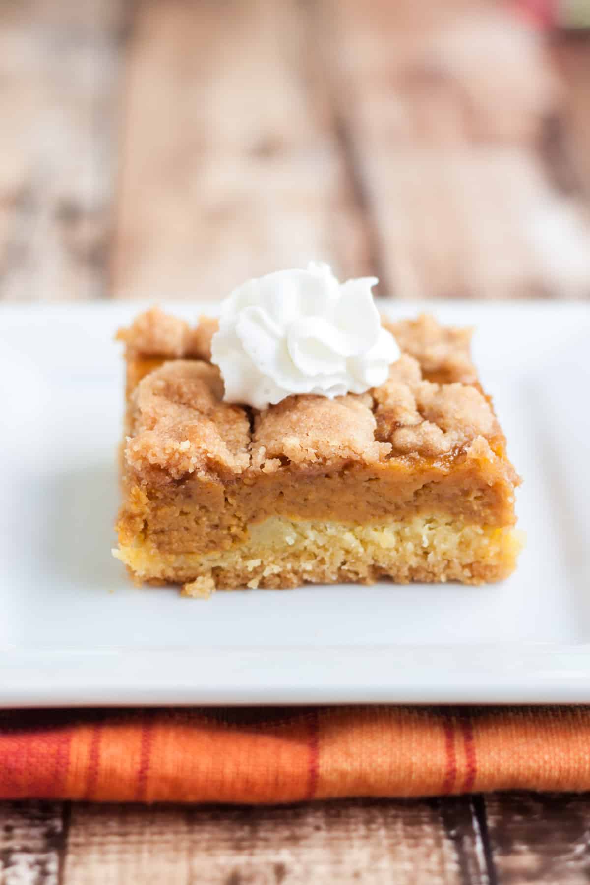 Pumpkin Pie Bar dessert with dollop of whip cream on top on a white plate with orange plaid napkin below.