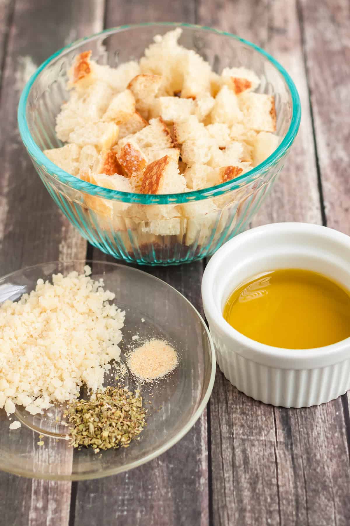 bread cubes in bowl, olive oil in small white bowl and grated parmesan cheese, dried basil and garlic powder on clear plate